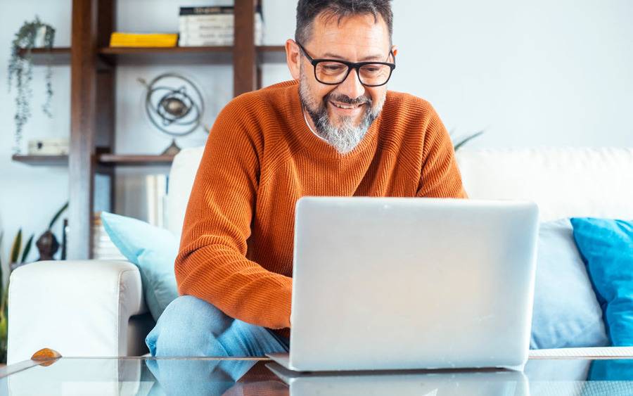 A man with gray hair smiles as he looks at a laptop while participating in a virtual support group for cancer survivors.