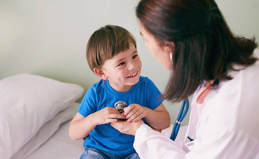 A pediatric patient in a blue shirt smiling at the provider caring for home while holding a stethoscope in his hands.