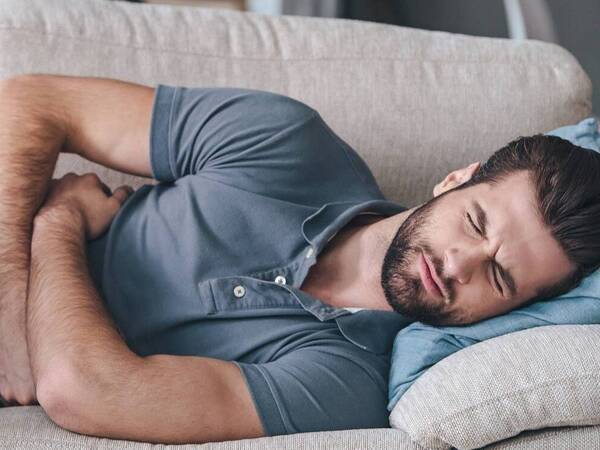 A young white man lies on the couch with his arms wrapped around his stomach and his eyes closed in pain.