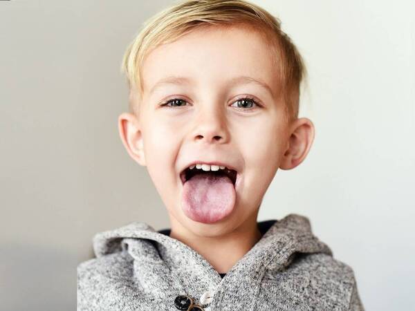 A young boy with blond hair sticks out his tongue to have his tonsils checked.