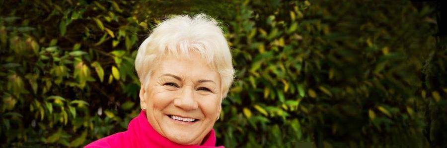 A cheerful elderly woman smiles while standing before a garden hedge.