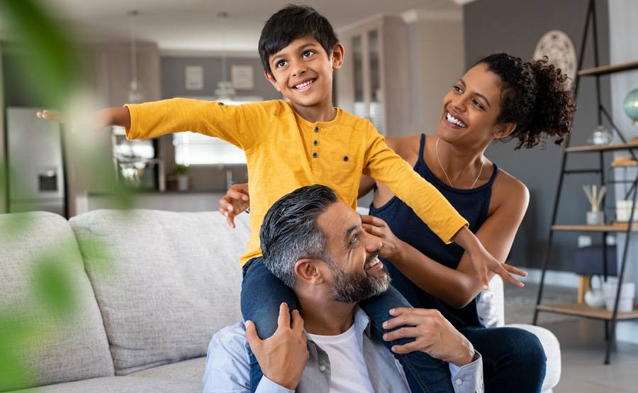 A young child sits on his father's shoulders in a living room, while his mom stands ready to catch him.