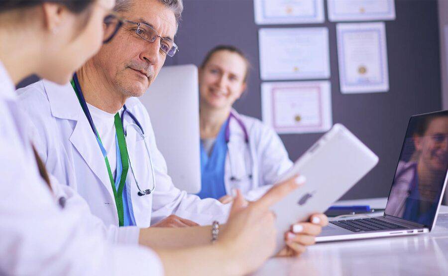 A group of physicians sits at a table with tablets and laptops, representing medical education and training programs.