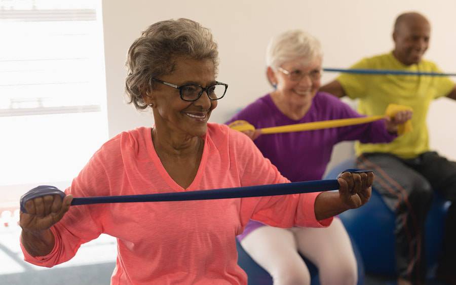A diverse group of older men and women smile while they participate in an exercise class using bands to improve their strength.