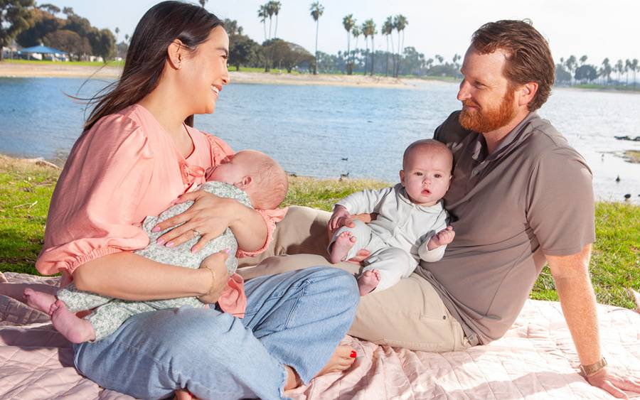Caitlin Welsh and her husband, Kevin, smile as they sit on a blanket by the bay each holding one of their twin babies.