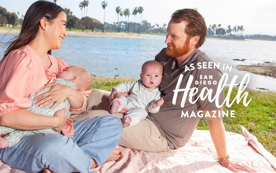 Caitlin Welsh and her husband, Kevin, smile as they sit on a blanket by the bay each holding one of their twin babies. San Diego Health Magazine