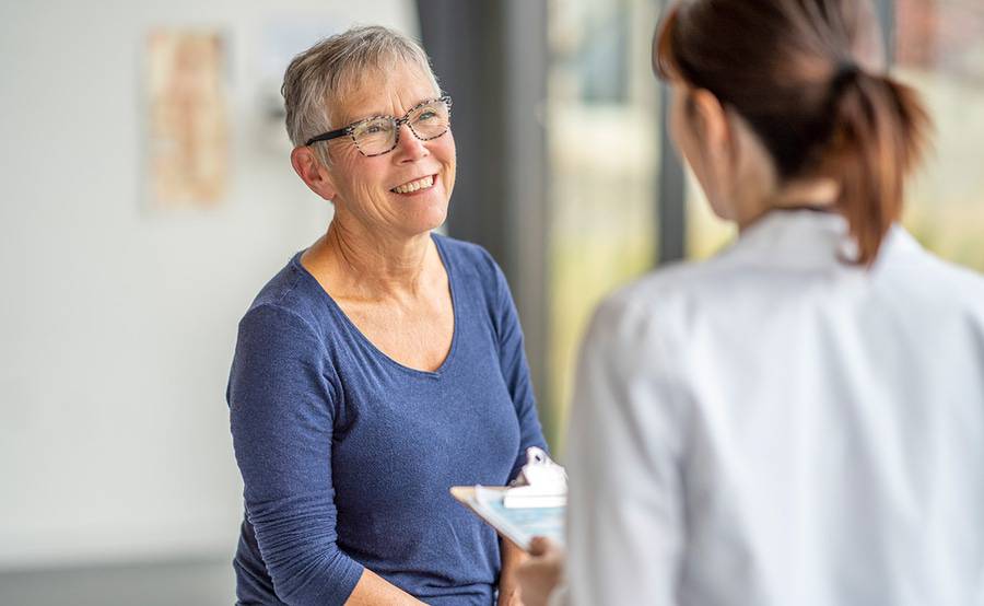 A mature woman wearing glasses is smiling and speaking with a health care provider.