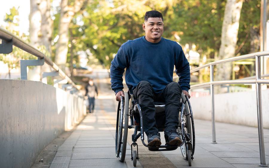 A young man in a wheelchair smiles as pushes himself up an outdoor ramp.