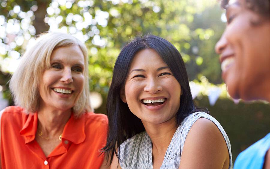 Three mature woman smile while sitting outdoors, representing an emotional support group for breast cancer.