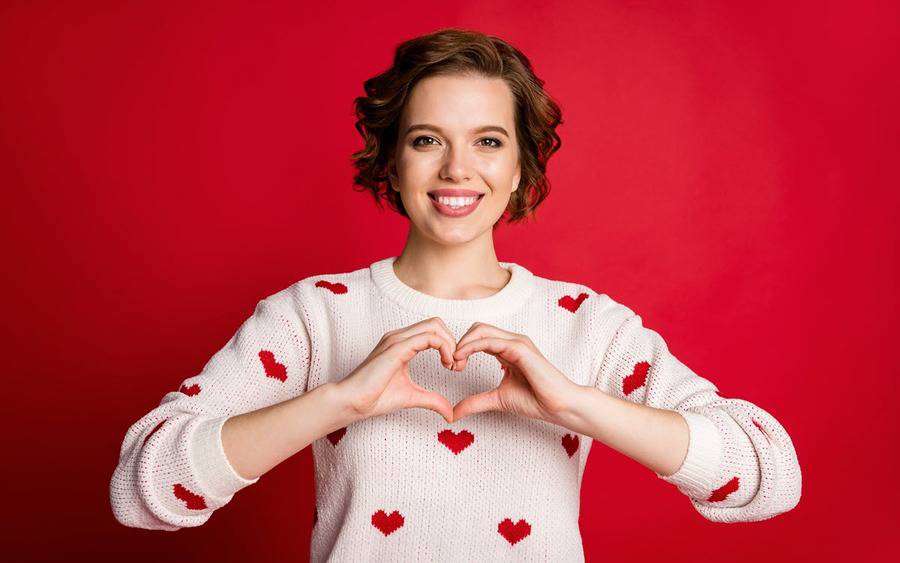 A woman smiles wearing a sweater with hearts. She makes a heart symbol with her hands in front of her heart.
