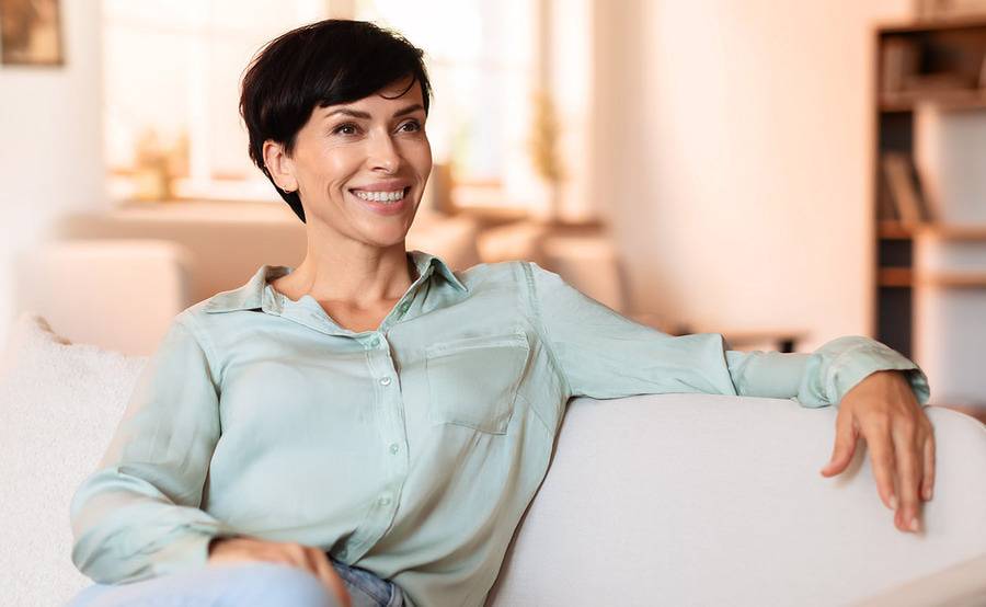 A woman sitting on a couch smiling in a well lit living room illustrating the quality of life with oculoplastic surgery at Scripps Health.